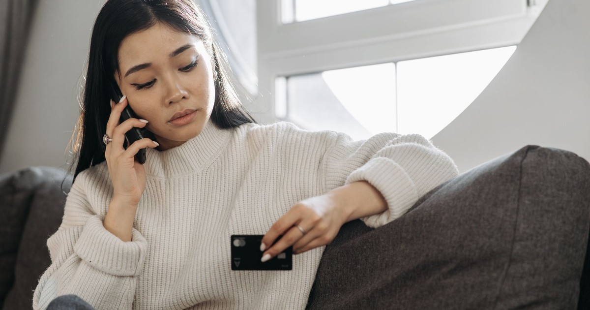 A woman sits on a sofa, multitasking with a phone and credit card, focusing on online shopping.