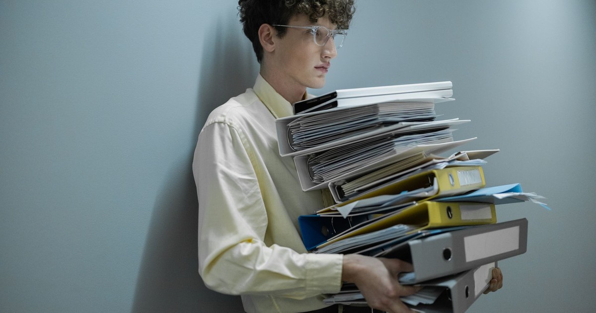 An overworked office worker carrying a large stack of files and folders in a modern setting.