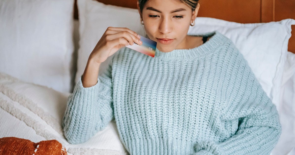 Crop concentrated Asian female in knitted blue sweater lying on cozy bed with credit card