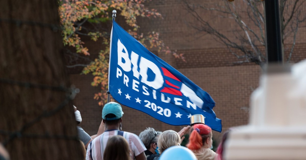 A diverse crowd gathered outdoors holding a Biden 2020 flag during a protest.