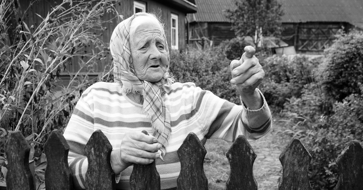 Black and white photo of an elderly woman in a rural setting, gesturing behind a wooden fence.