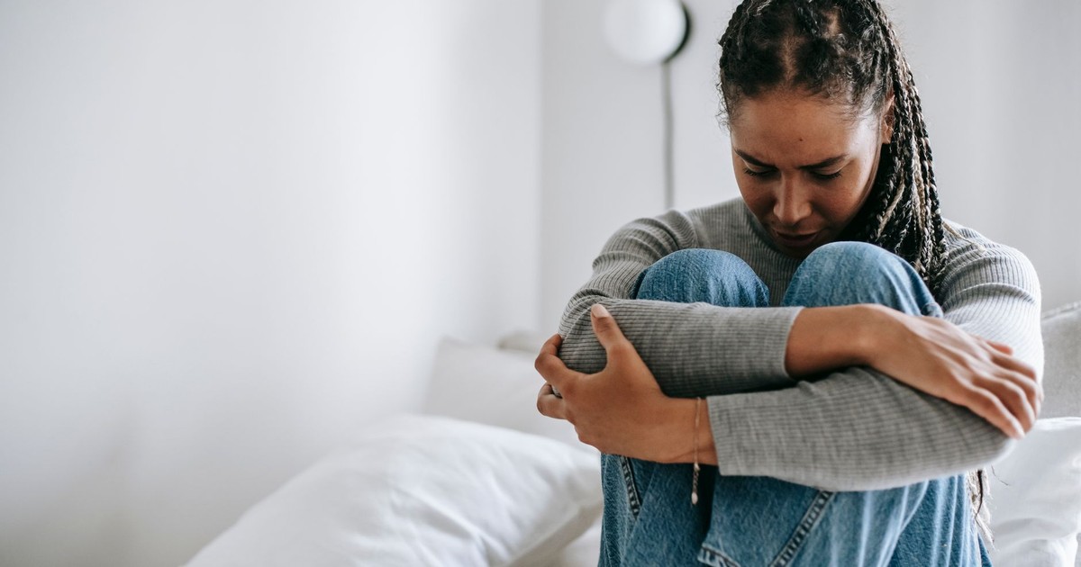 Disappointed ethnic female embracing legs while suffering from problem and sitting on white bed