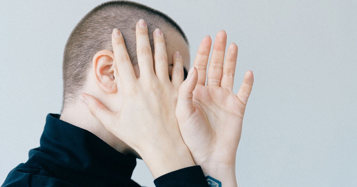 A person hides their face with hands in a minimalist portrait with a light background.