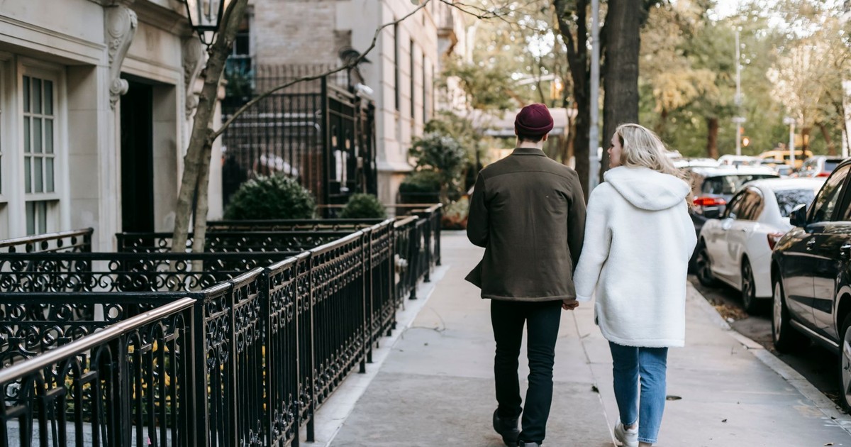 Back view of anonymous trendy young couple in stylish warm clothes holding hands and walking in city street near residential houses in autumn
