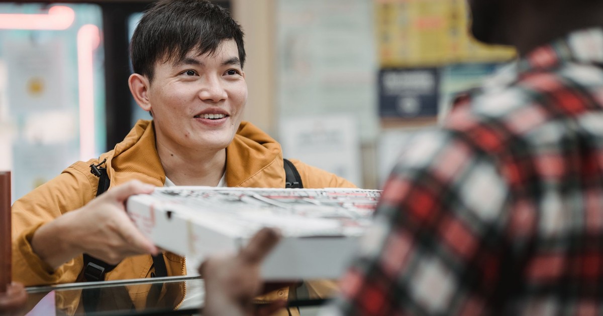 A cheerful Asian man receives a pizza box inside a shop, engaging with the seller.