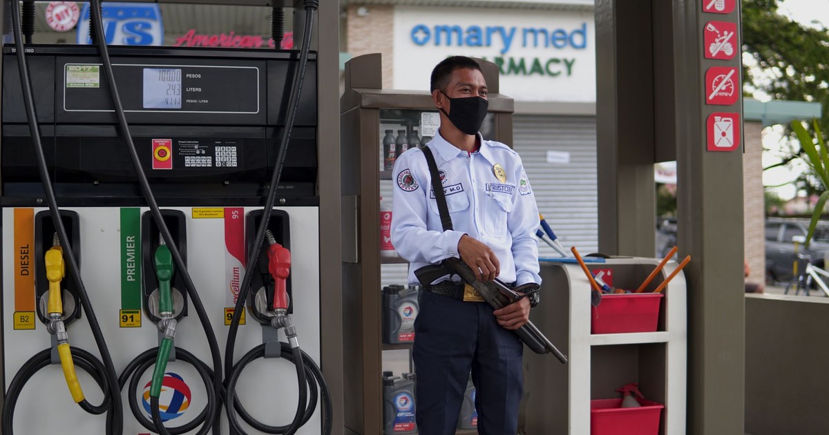 A uniformed security guard wearing a face mask stands vigilant at a gasoline station with fuel pumps.