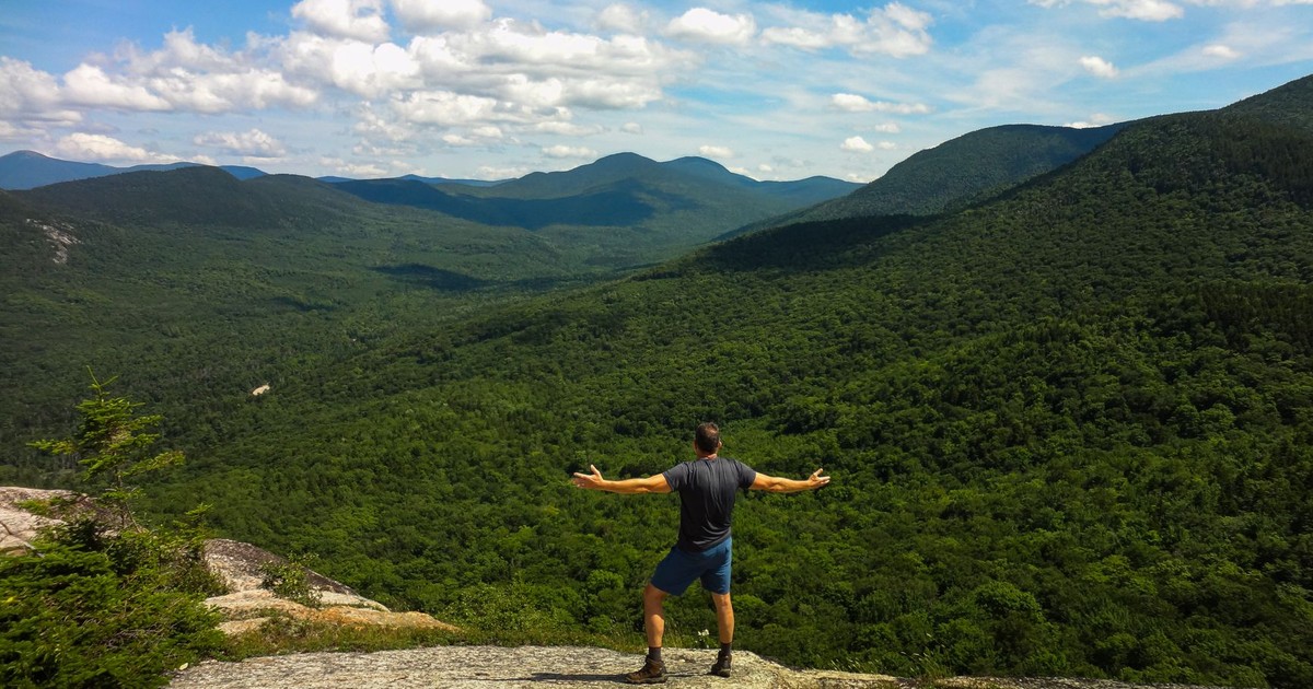 Hiker embracing nature on a rocky cliff, overlooking lush valleys in White Mountains, NH.
