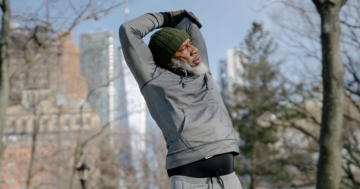 Senior African American male in hoodie and cap raising arms during training in park and looking away