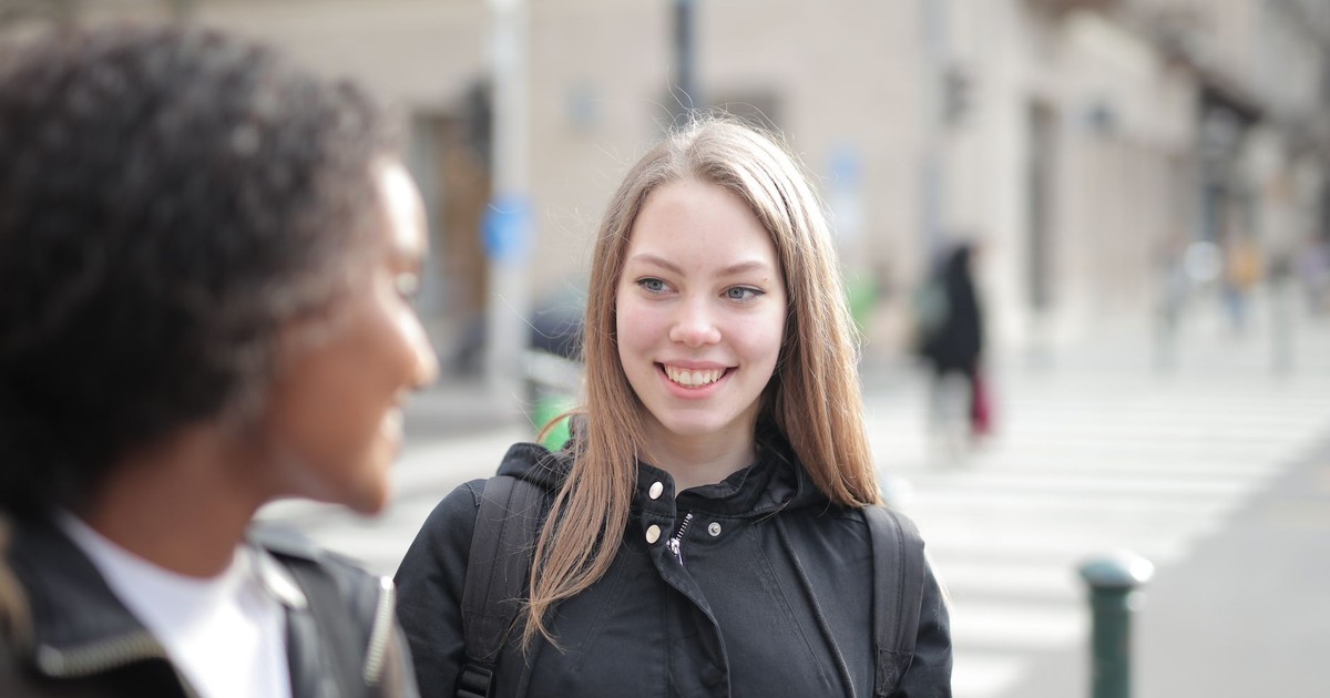 Two young women smiling and talking on a city street during the day.