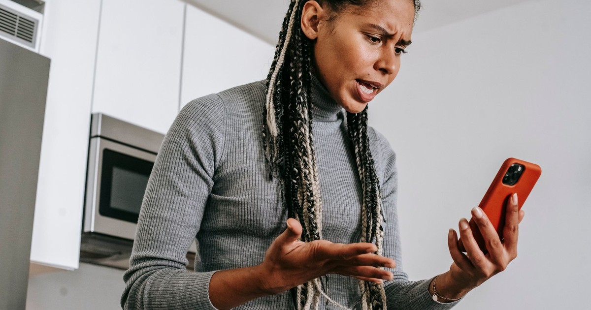 From below of angered black female with Afro braids in casual outfit gesticulating and resenting while having video call on smartphone in modern light kitchen