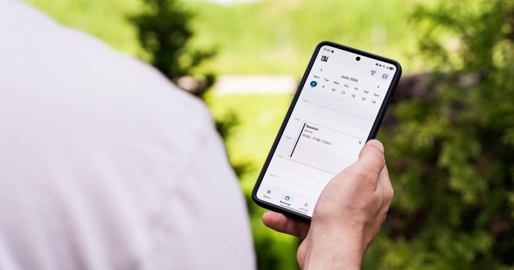 A person using a sauna booking app on a smartphone outdoors, surrounded by greenery.