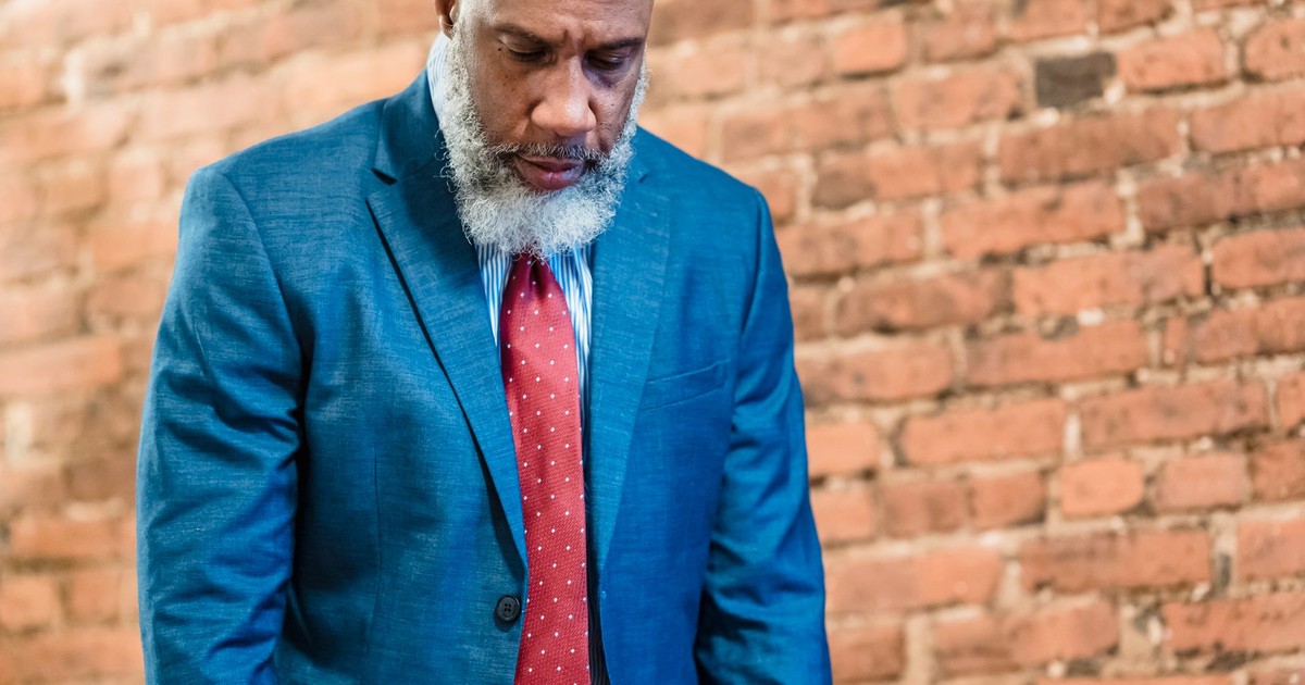 Elderly man in formal attire, appearing thoughtful against a brick wall backdrop.