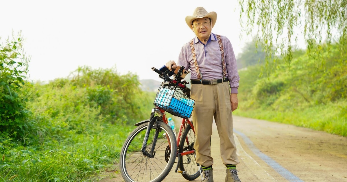 Elderly man standing with a bicycle on a rural path, wearing suspenders and a cowboy hat.