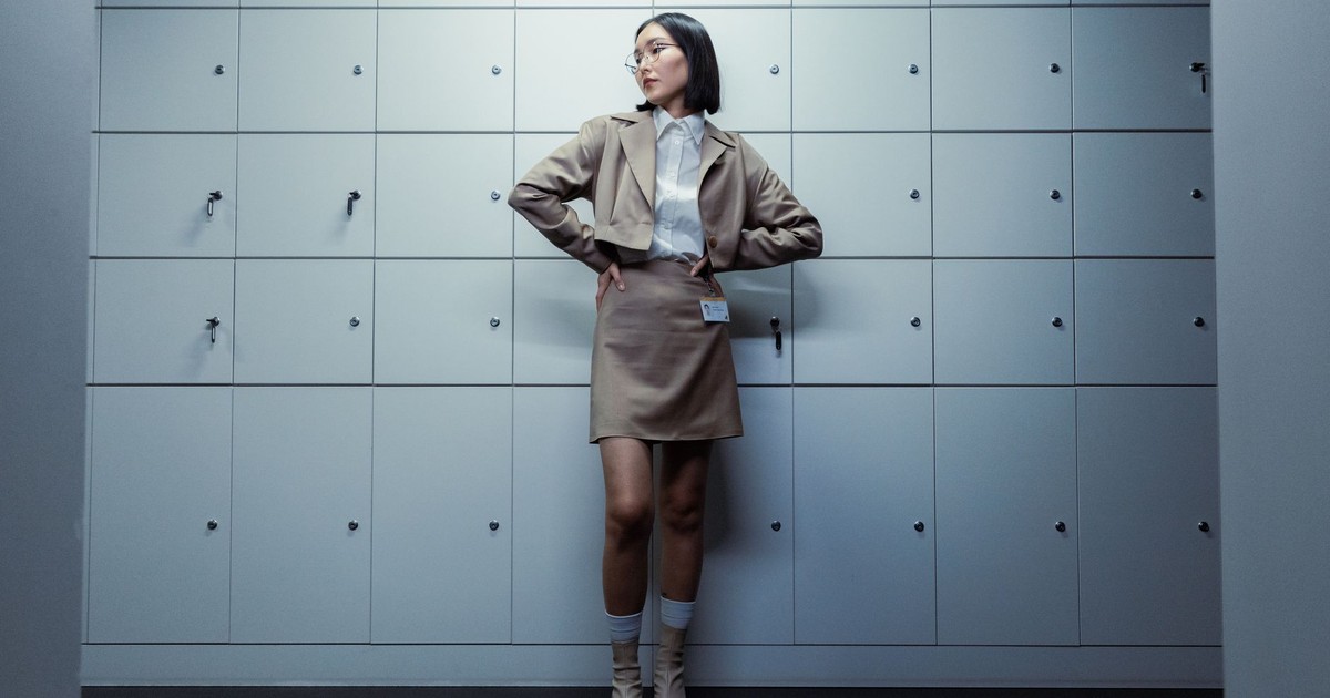 Confident woman in a blazer and skirt posing in front of office lockers, looking aside.
