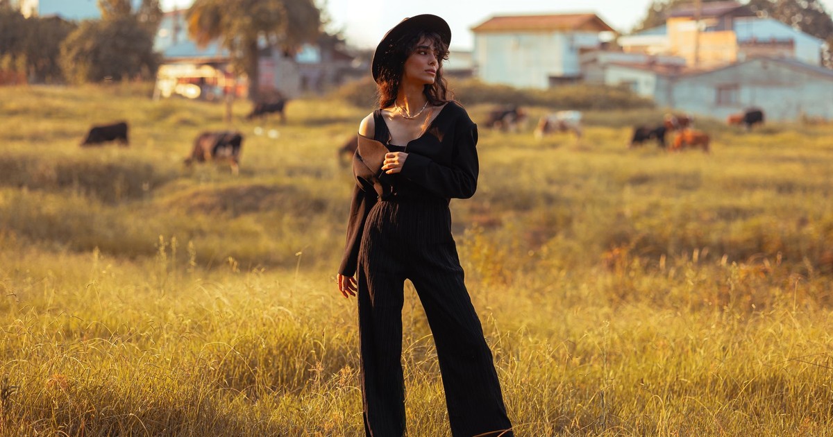 Woman in black outfit and hat in a rural field with cows in the background.