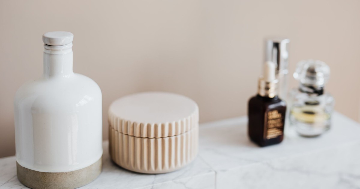 Set of reusable cosmetic containers for cream and shampoo arranged near various skincare products and perfume bottle on white marble shelf against beige wall in modern bathroom interior