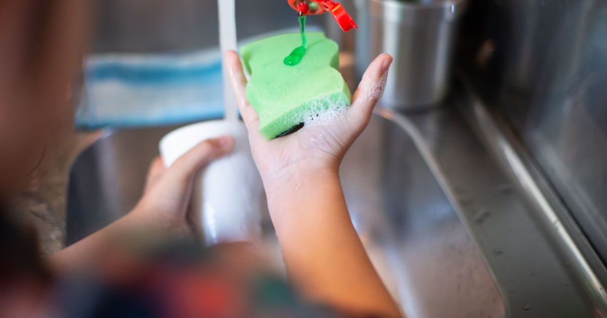 Hands pouring liquid soap onto a sponge under running water in a kitchen sink.