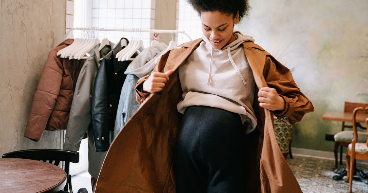 Pregnant woman in stylish coat adjusting outfit indoors near clothes rack.