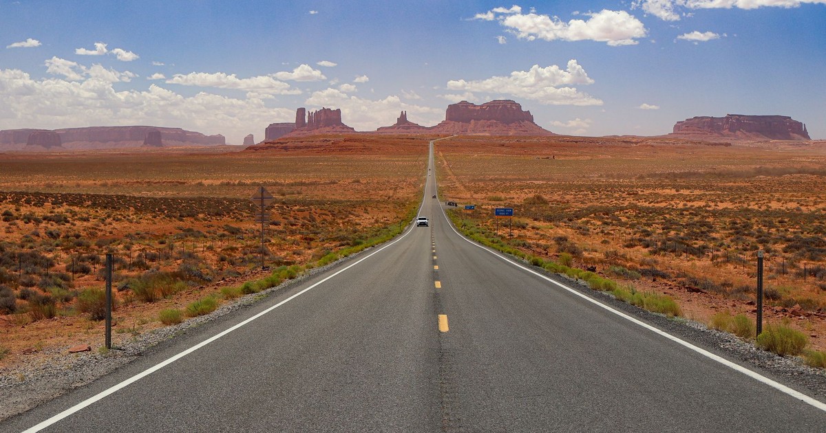 A long, empty highway stretches through Monument Valley under a bright blue sky.