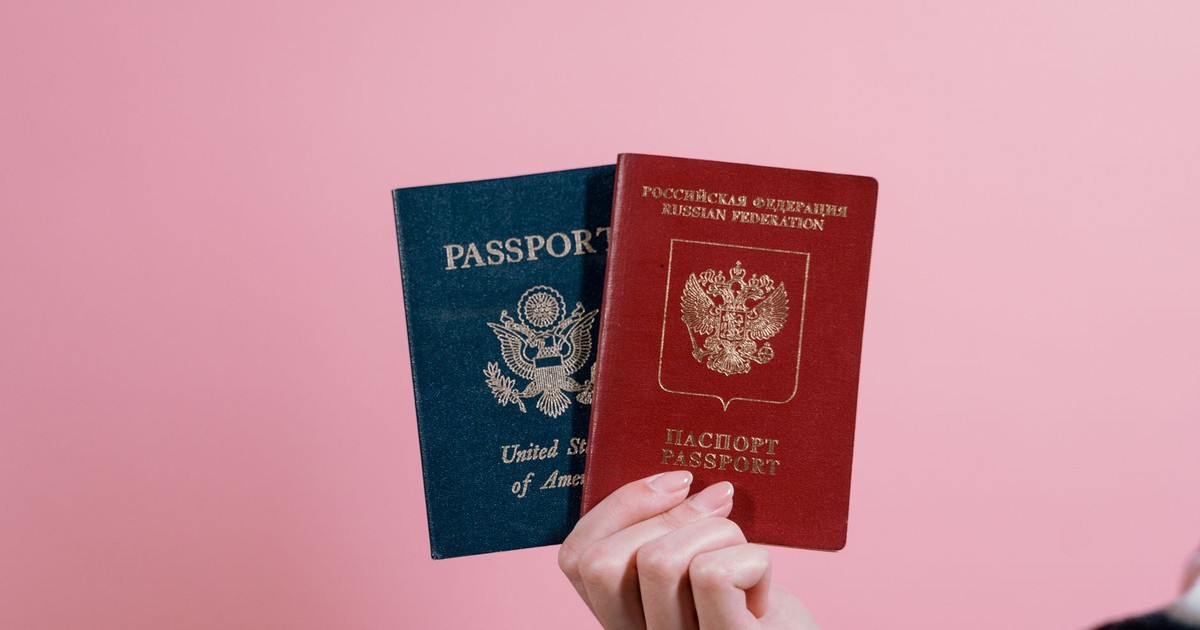 Close-up of a hand holding American and Russian passports on a pink background.