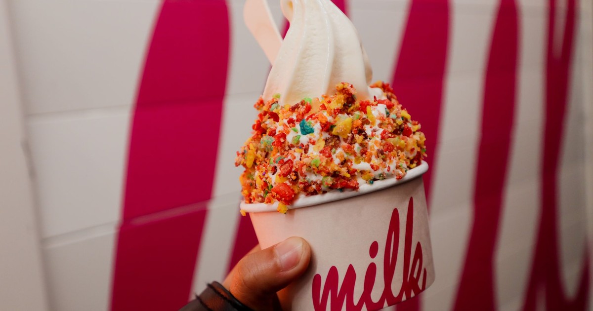 Hand holding a vanilla soft serve ice cream with colorful toppings in a vibrant setting.