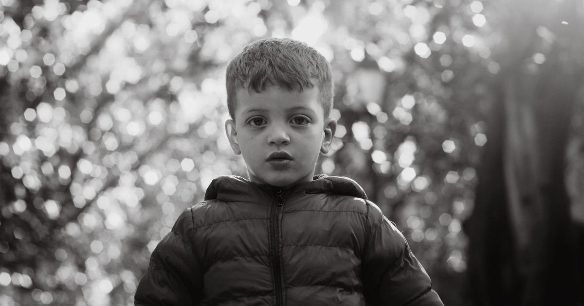 A striking black and white portrait of a young boy wearing a jacket, outdoors.