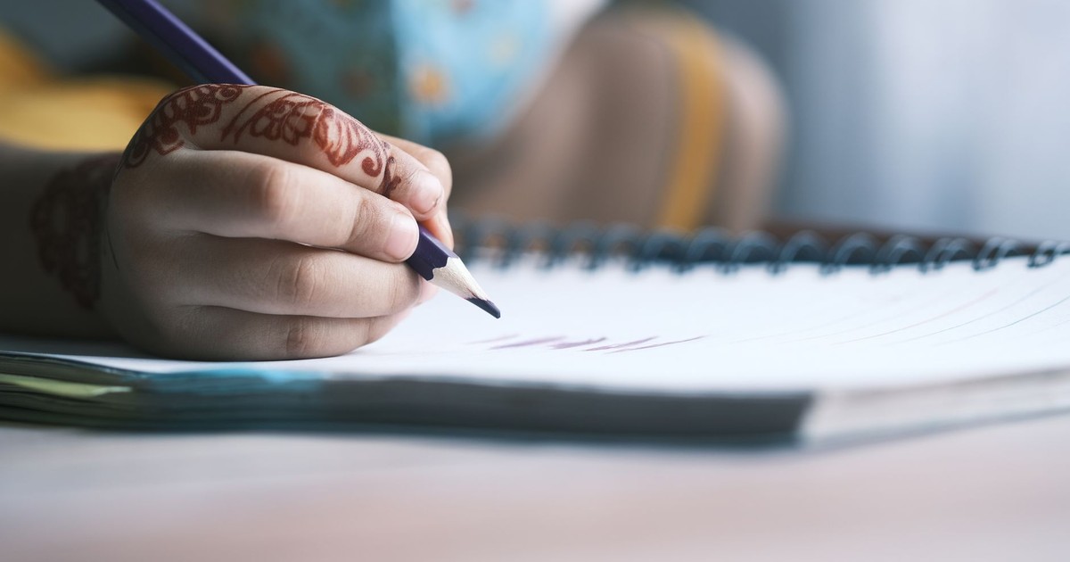 Close-up of a child's hand writing in a notebook with colorful pencils and henna design.