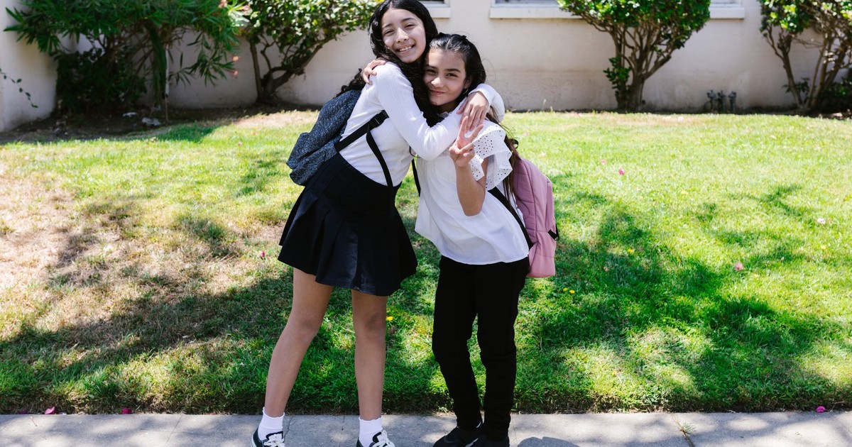 Two friends hugging outside a school building on a sunny day, both wearing backpacks.