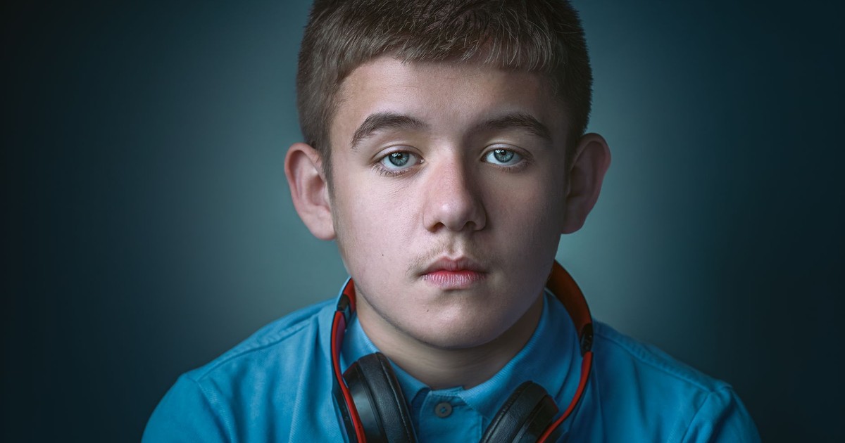 Portrait of a serious teenage boy wearing a blue blouse and headphones against a gray background.