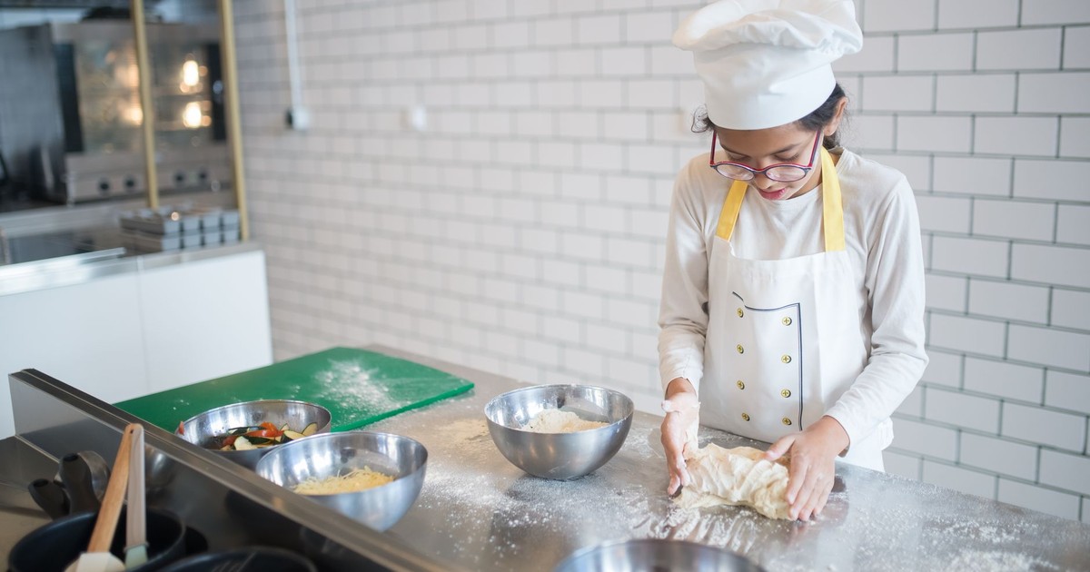 A young girl in a chef's hat kneads dough in a contemporary kitchen setting.