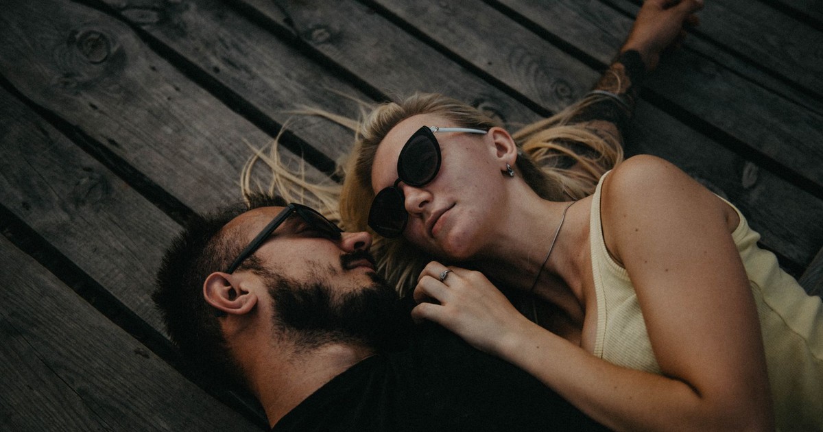A couple laying on a wooden floor, embracing in a romantic moment with sunglasses on.