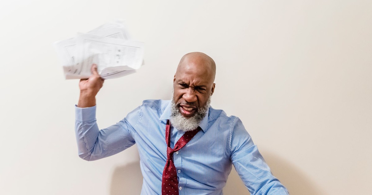 A frustrated bald man with a beard throws papers in anger, expressing intense emotion.