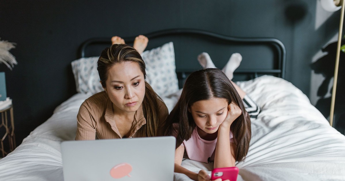 Mother and daughter lying on bed using laptop and smartphone indoors, depicting modern family life.