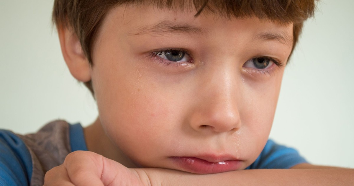 Portrait of crop upset little boy with brown hair and green eyes crying and resting head on arm while looking away against white background