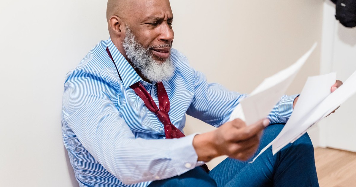 An adult man with a beard looks distressed while sitting on the floor, examining papers.