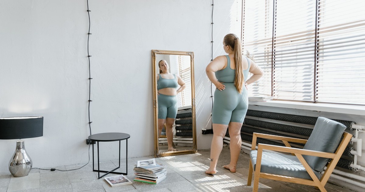 Plus size woman in sportswear stands barefoot, looking at her reflection in a wall mirror.
