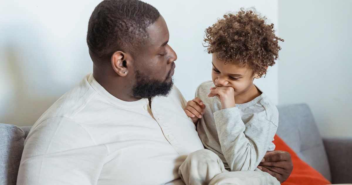 Calm African American father looking at cute black son sitting on knees on couch near wall in light room at home