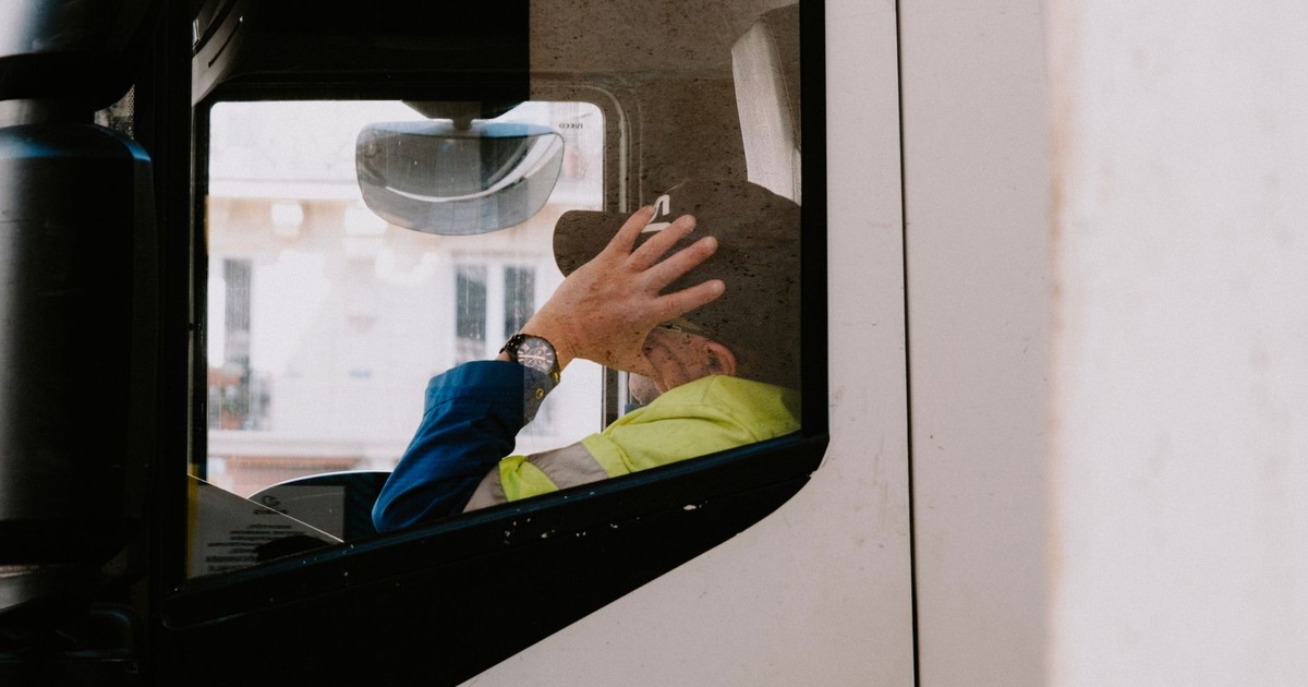 A truck driver in neon jacket rests with hand on head inside vehicle cabin, view from the side window.