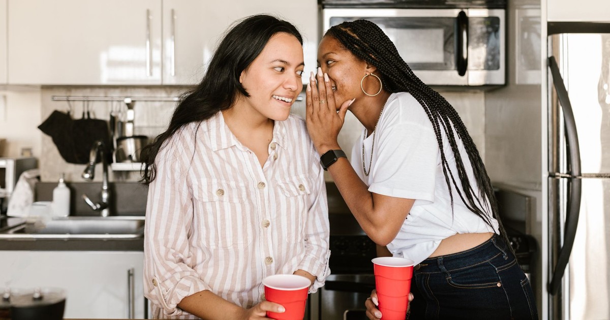 Two diverse women enjoying a candid moment and whispering secrets in a modern kitchen setting.