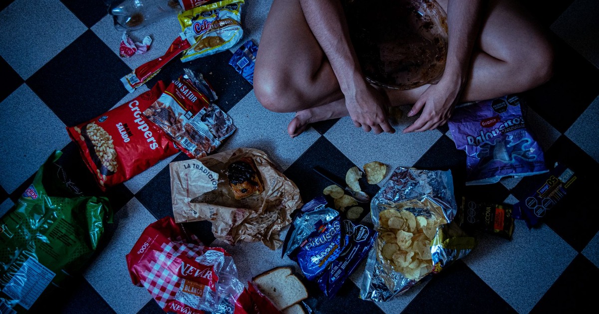 Man sitting surrounded by assorted snacks and packaging on a checkered floor, depicting a binge-eating scene.