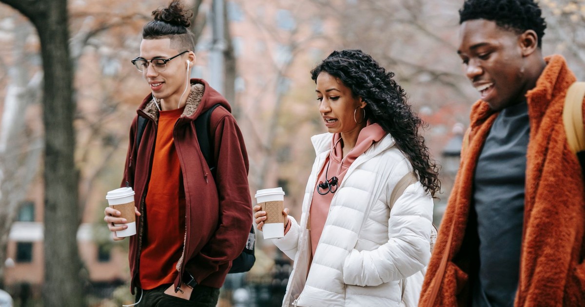 Confident young multiracial friends in outerwear and backpacks talking while strolling in city street near trees with paper cups with drink and earphones in daytime in park