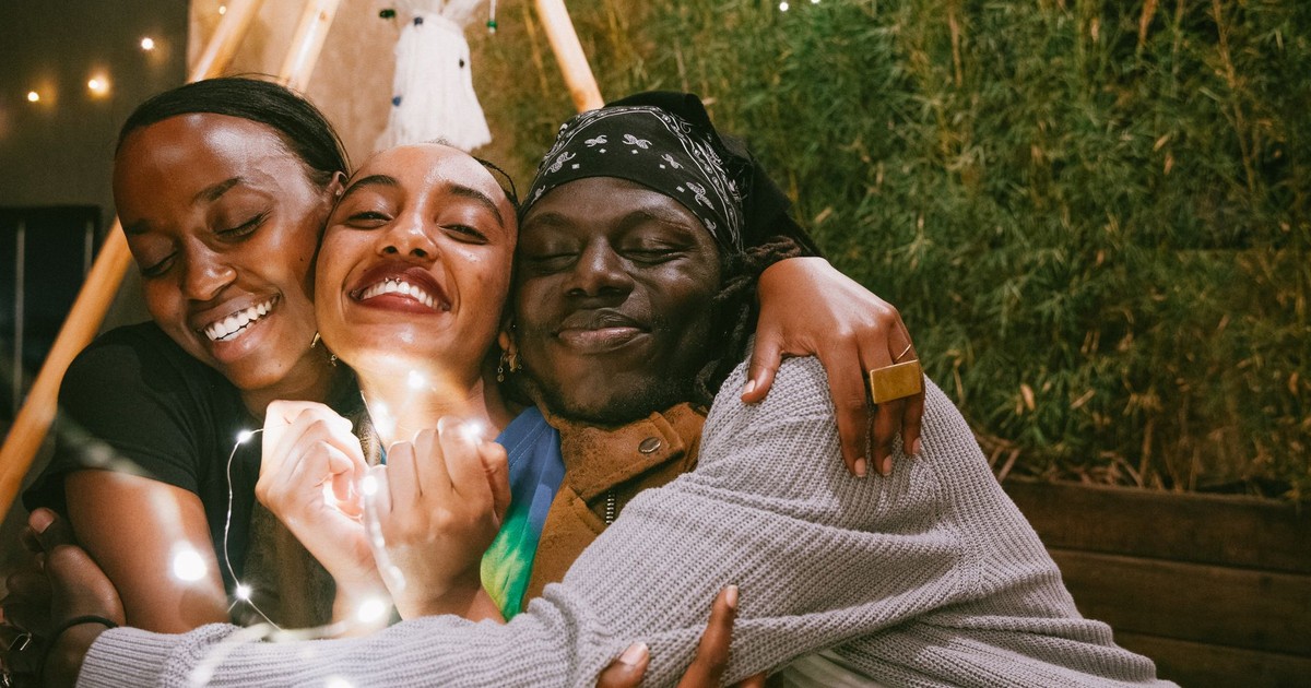 Three friends share a joyful embrace, radiating happiness under string lights.