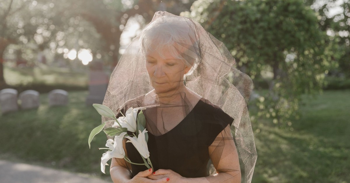 Senior woman in black dress and veil holding white lilies in a cemetery.
