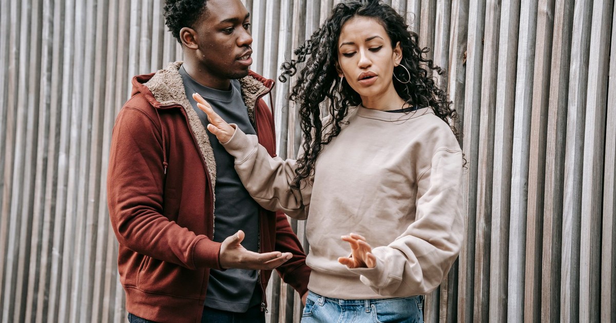 Annoyed ethnic girlfriend with curly hair pushing away concerned African American boyfriend while having conflict on street near wooden wall during breakup