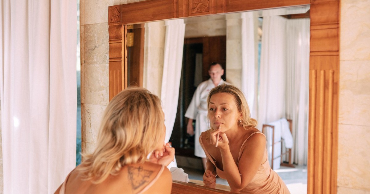 A woman applies makeup in a sunlit bathroom with warm, natural tones.