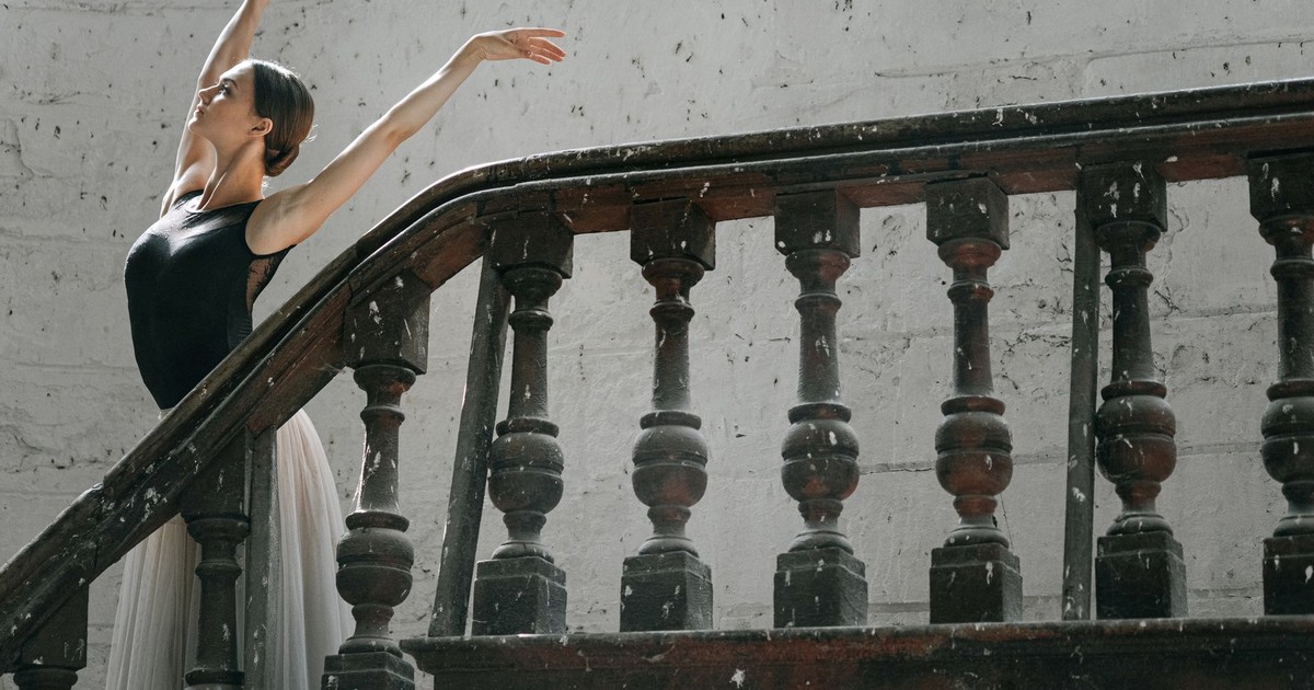 A graceful ballerina poses elegantly with hands raised beside a vintage staircase.