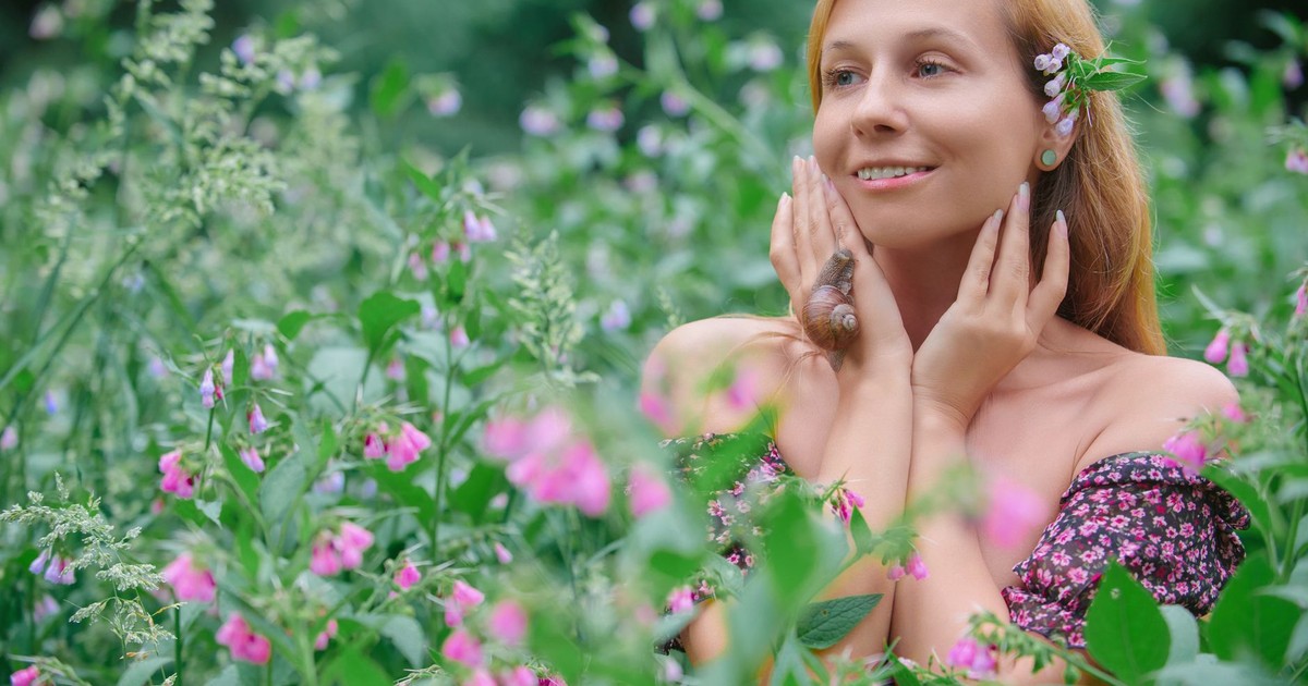 Smiling woman surrounded by pink flowers in lush greenery, symbolizing nature's beauty.