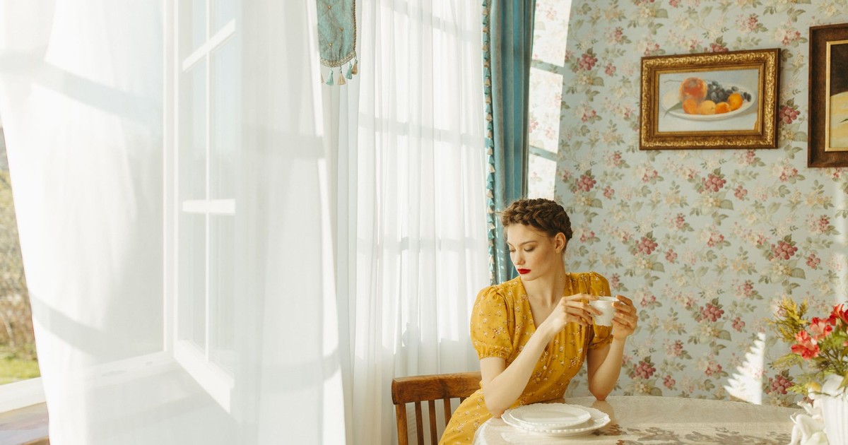 A woman in a yellow dress enjoys coffee by a sunlit window in a vintage room.
