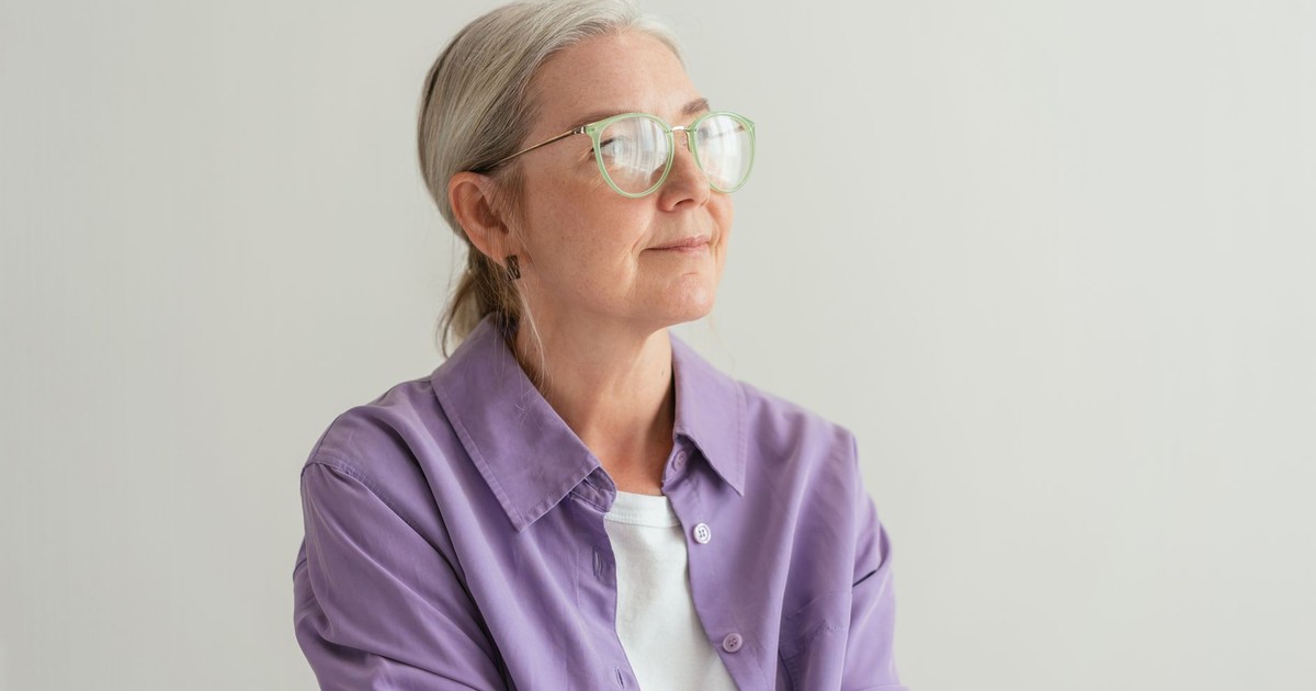 Senior woman with grey hair wearing eyeglasses and a purple shirt, looking thoughtful.