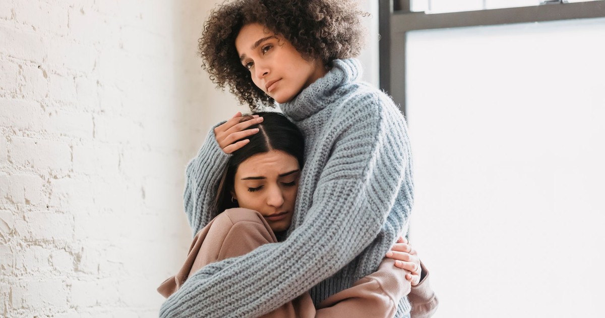 Frustrated African American woman embracing and comforting sad female friend while standing near window and wall in room at home